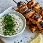 Grilled Chicken Bites with Creamy Garlic Dipping Sauce Handheld smartphone photo of grilled chicken skewers with distinct dark char marks and a bowl of opaque white garlic sauce. (Grilled Chicken Bites)