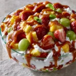Eye-level close-up of a Cheesecake Fruit Salad in a glass bowl with visible layers of whipped cream, red strawberries, and golden caramel sauce.