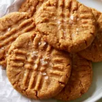 Oat Flour Peanut Butter Cookies with Flaky Sea Salt High-angle shot of golden-brown Oat Flour Peanut Butter Cookies stacked on a white ceramic plate with visible rustic cracked edges.