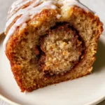 Cinnamon Roll Banana Bread: The Ultimate Moist Swirl Loaf with Thick Glaze A thick, moist slice of golden-brown Cinnamon Roll Banana Bread on an off-white plate featuring a prominent dark brown cinnamon-sugar swirl.
