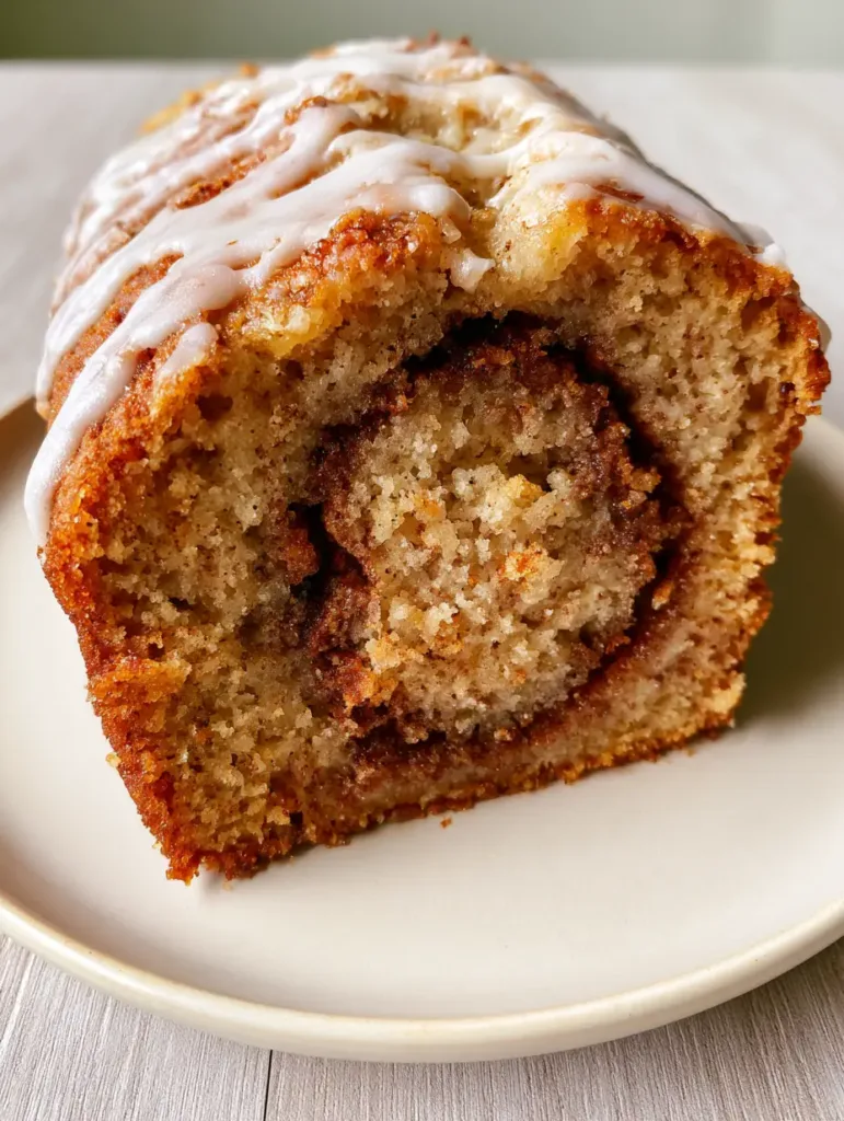 A thick, moist slice of golden-brown Cinnamon Roll Banana Bread on an off-white plate featuring a prominent dark brown cinnamon-sugar swirl.