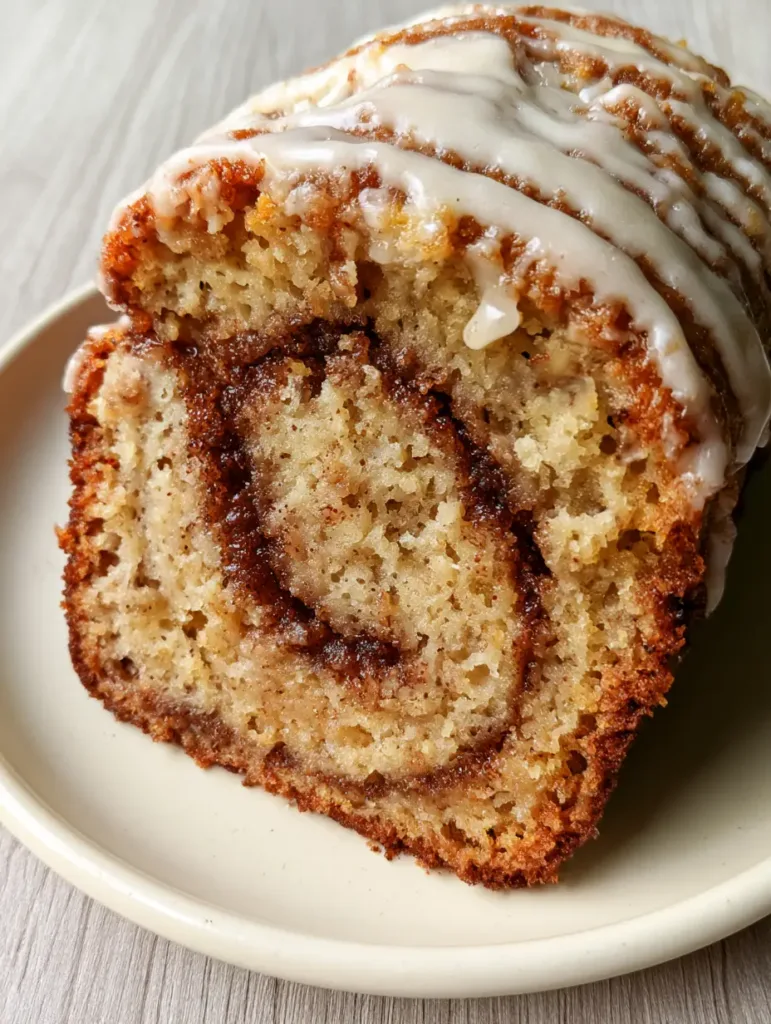 A slice of Cinnamon Roll Banana Bread on a wooden surface, highlighted by soft natural light showing the thick white glaze drips.