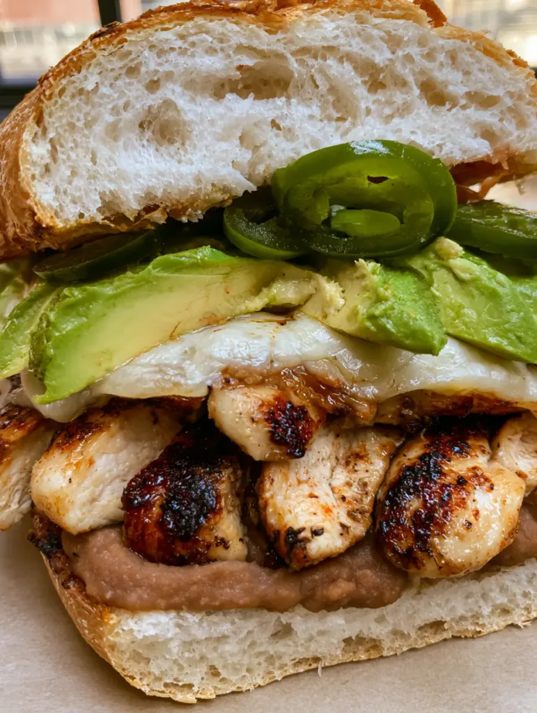 A close-up of a Chicken Torta Sandwich showing deep brown sear marks on chicken and melting white Oaxaca cheese.
